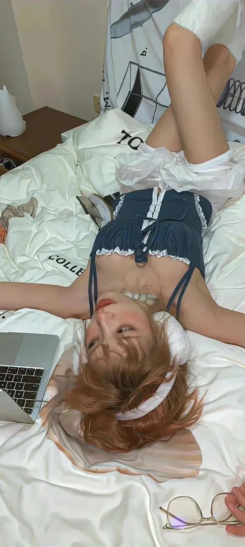 金发少女躺卧在卧室白色床品上，身旁摆放笔记本电脑，姿态慵懒放松