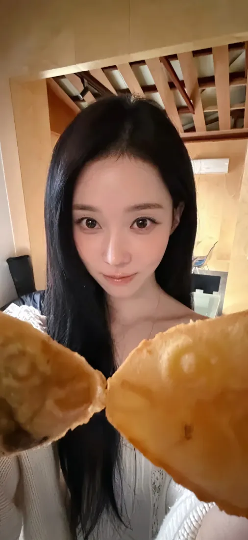 室内自拍，黑长直长发的金玟庭手持两块面包凑近镜头，背景为暖调木质室内空间。