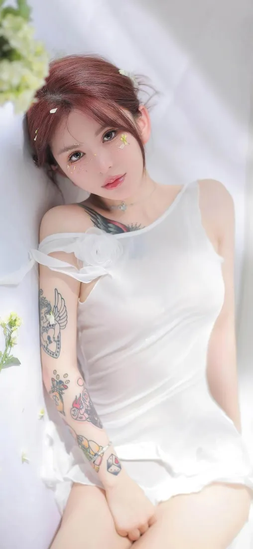 红发花臂美女身着白色吊带裙，倚靠在带绿植的白色简约背景前。