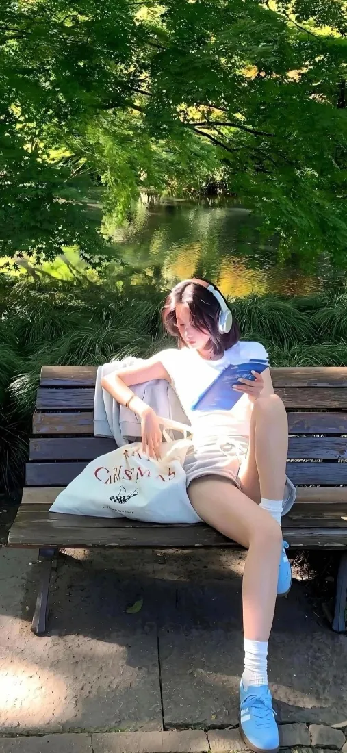 戴白色耳机的女生坐在公园木质长椅上，手持手机，身旁放着白色布袋，背景是茂密绿植。