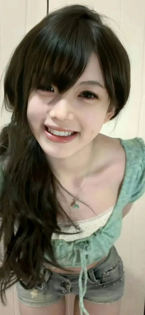 室内浅色墙前，黑发长卷发穿绿上衣的年轻女生露齿甜笑自拍。