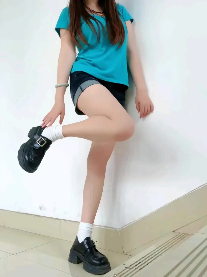 长发女生靠墙站立，穿蓝色短袖黑色短裤，单腿抬起展示厚底皮鞋