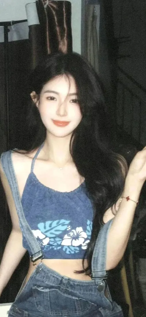 夜晚室内，黑发长发年轻女子穿蓝色印花吊带与牛仔背带裤，微笑看向镜头。
