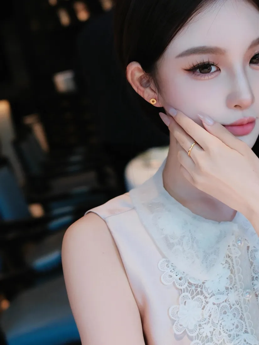 夜晚暗调虚化背景中，穿白色蕾丝无袖裙的女生手托脸颊，佩戴金色耳钉与戒指。