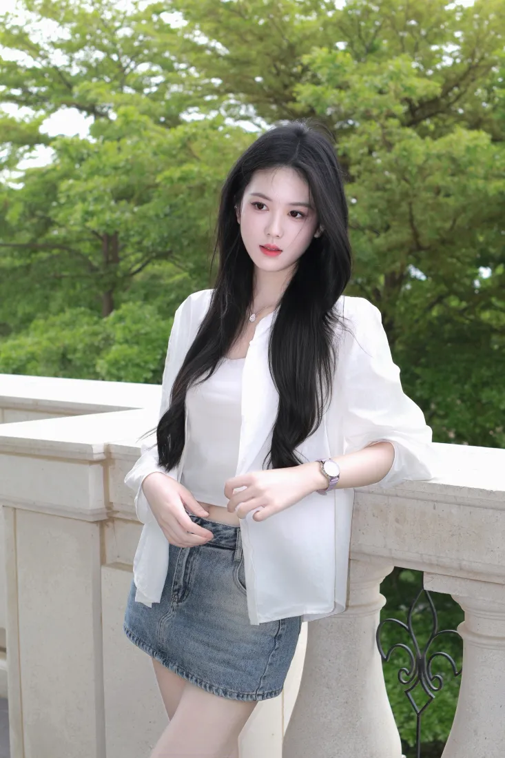 户外绿树旁，长发白衣牛仔短裙美女倚石栏杆休闲站立
