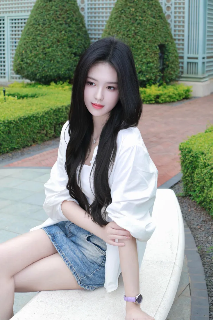 户外园林中，长发白衣牛仔短裙女生坐在白色弧形石凳上看向镜头