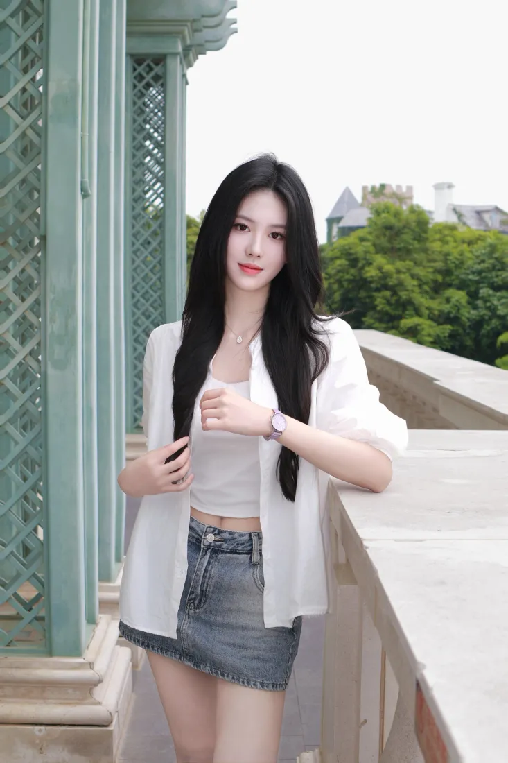 长发年轻女子身着白色开衫与牛仔短裙，倚靠在露台石质护栏旁，背景是绿树与远处建筑。