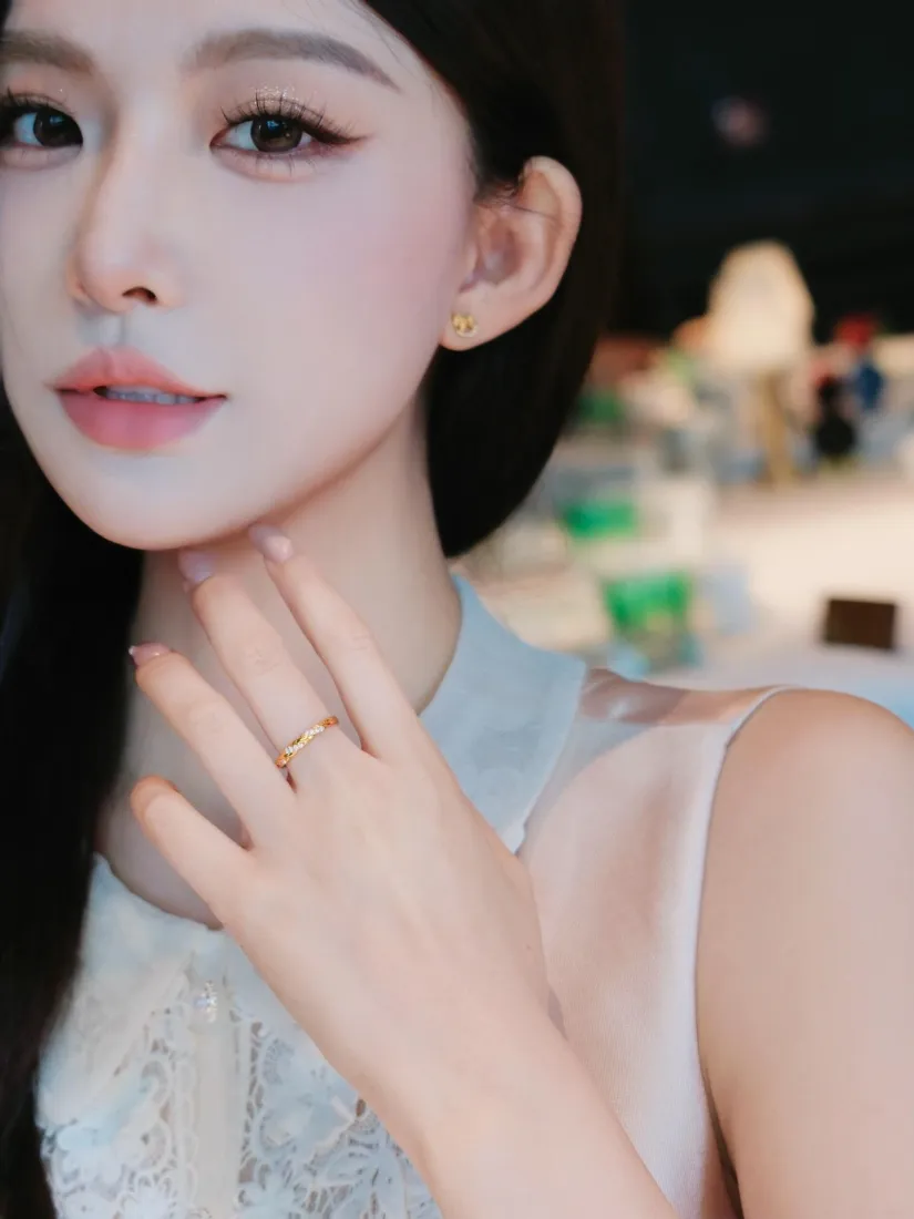 长发精致妆容的年轻女性，佩戴金耳钉与金戒指，手轻触脖颈，背景虚化。