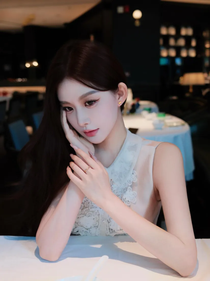 长发女子身着白色无袖上衣，手肘撑在餐厅桌面，手托脸颊看向镜头。