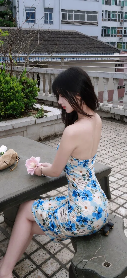 天台露背蓝碎花裙长发美女坐于石桌旁，手持粉花，背景为居民楼与绿植