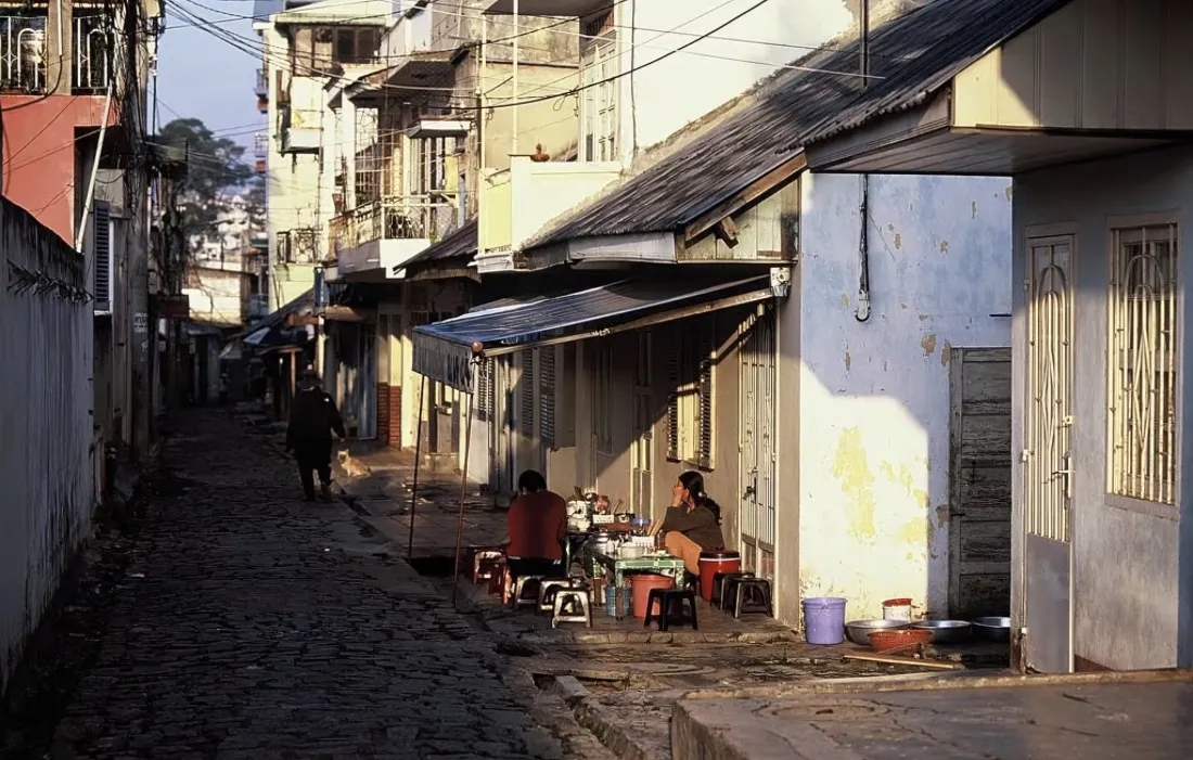 晨光下的老城石板小巷，两侧民居错落，居民闲坐路边，行人缓步走过。