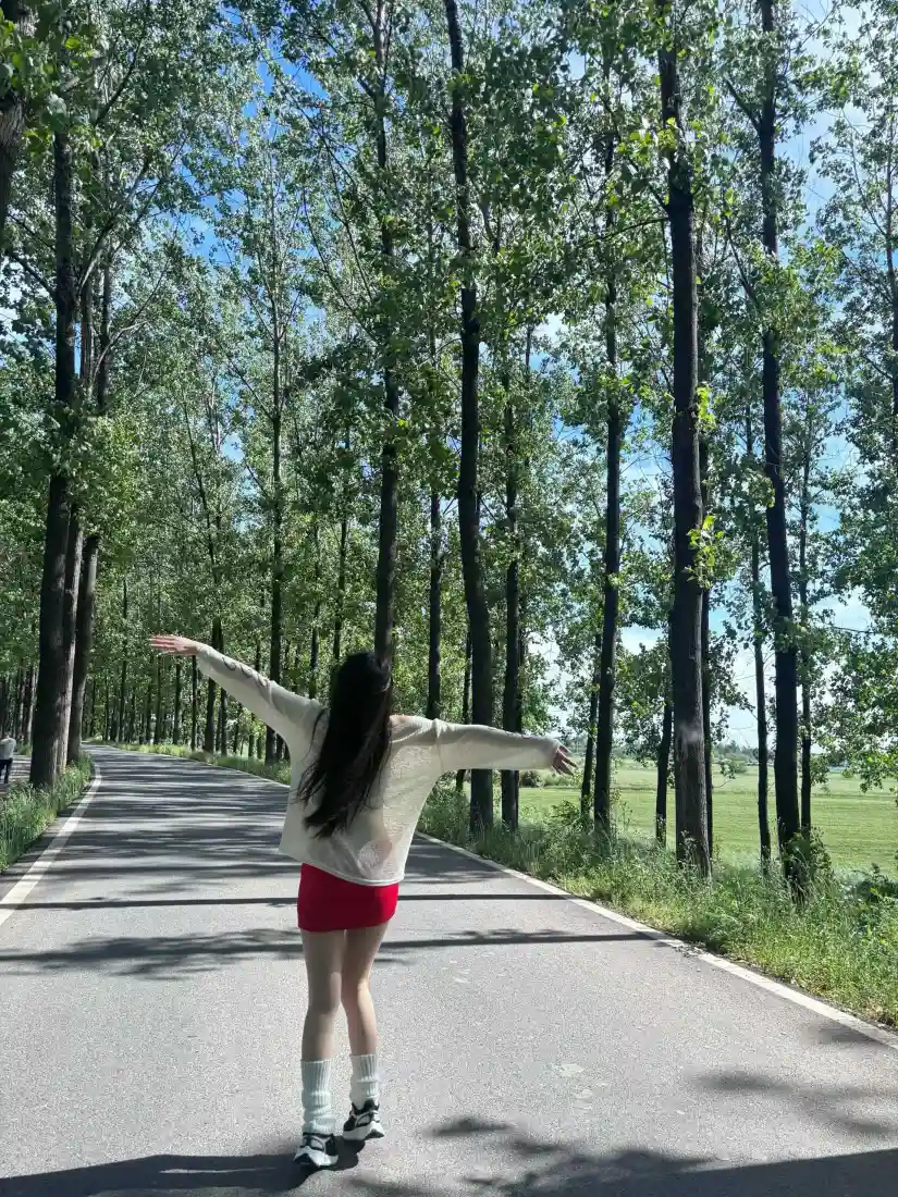 林间公路上，长发女生背对镜头张开双臂，两侧是高大绿树与蓝天。