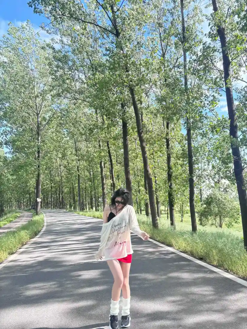 戴墨镜的年轻女子站在林间公路上，两侧是高大绿树与晴朗蓝天。