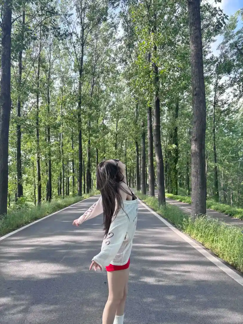 长发女生站在林间柏油路上，双臂舒展，两侧是高大绿树，阳光洒落。
