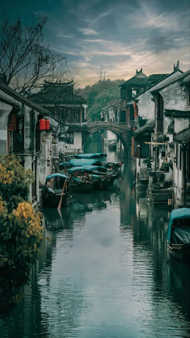 黄昏时分江南水乡河道，两岸白墙黑瓦古民居，乌篷船停泊，远处石拱桥横跨水面。