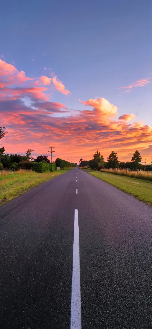 黄昏时分笔直延伸的郊外公路，两侧草地树木，天空布满橙粉晚霞