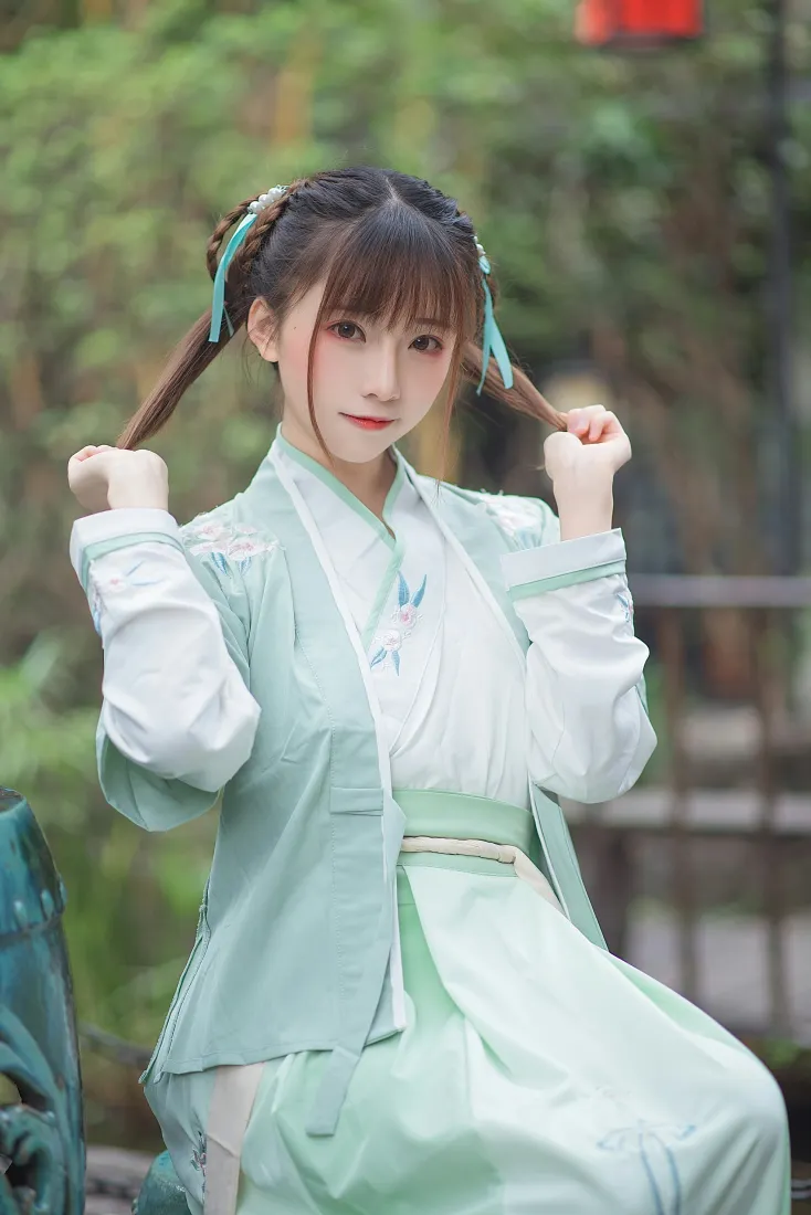 户外绿植背景前，穿淡绿汉服的双马尾少女双手轻抓发辫，神态甜美俏皮。