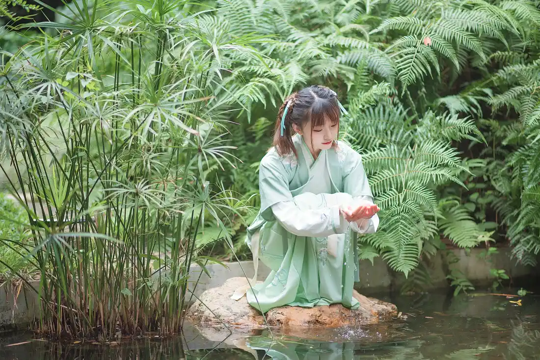身着浅绿色汉服的双马尾少女跪坐水边石块上，双手捧水，周围绿植环绕。