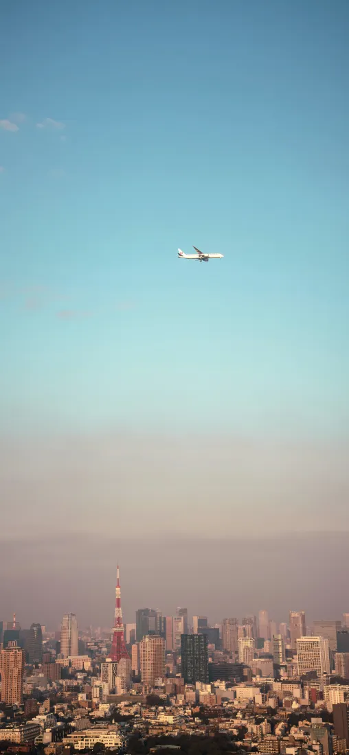 高空俯瞰东京城市天际线，东京塔矗立楼群间，民航客机飞过渐变蓝天