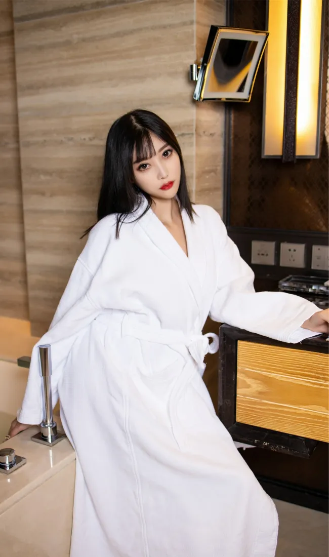 黑长直红唇女子身着白色浴袍，倚靠在酒店浴室台边。