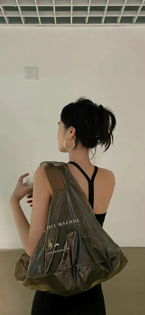 扎丸子头的女生背对镜头侧头，穿黑色吊带，挎棕色大包手持饮品，背景为素白室内墙面