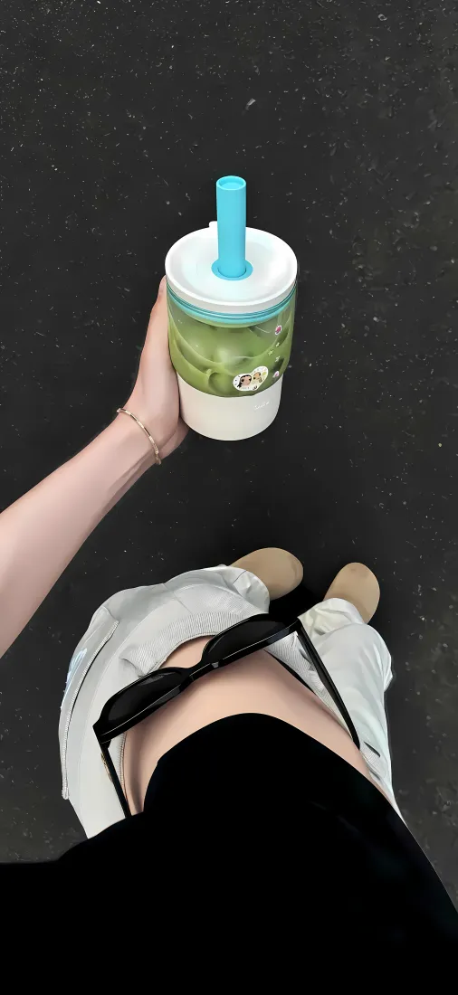 第一视角俯拍女生手持绿色冰饮，身着休闲短裤与小白鞋站在深色地面上