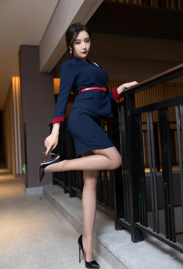 身着藏蓝职业套装配红腰带的女性在楼道扶栏抬腿摆拍，脚踩黑红细高跟