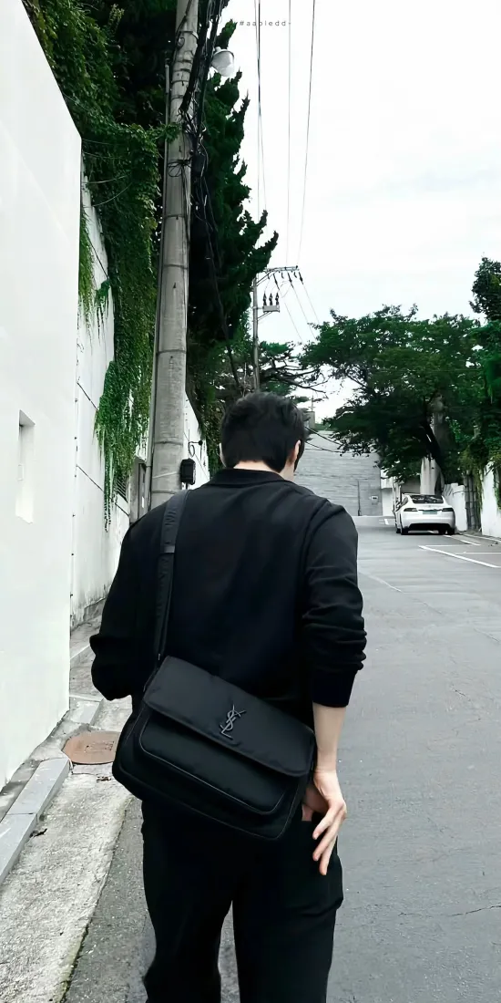 阴天街道上，黑衣斜挎包男生背对镜头向前行走，旁有白墙绿植。