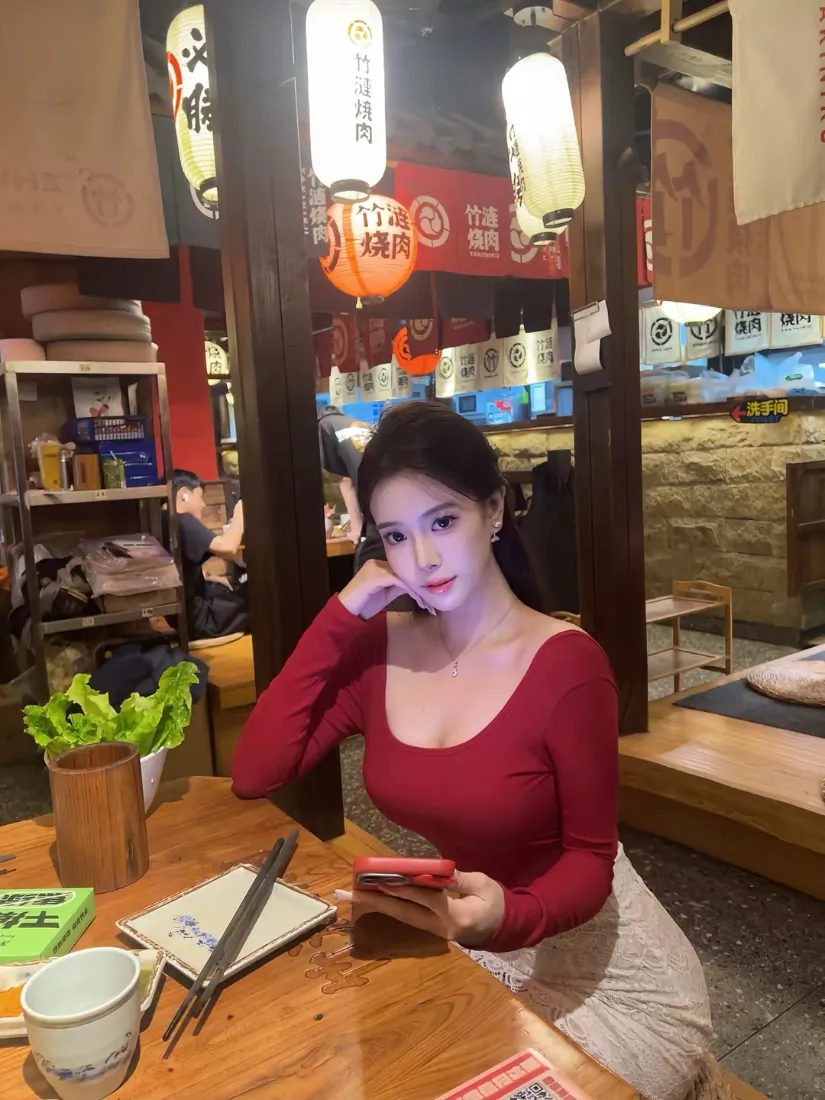 红衣长发女子坐在日式餐饮店木桌旁，手持手机，背景悬挂灯笼。