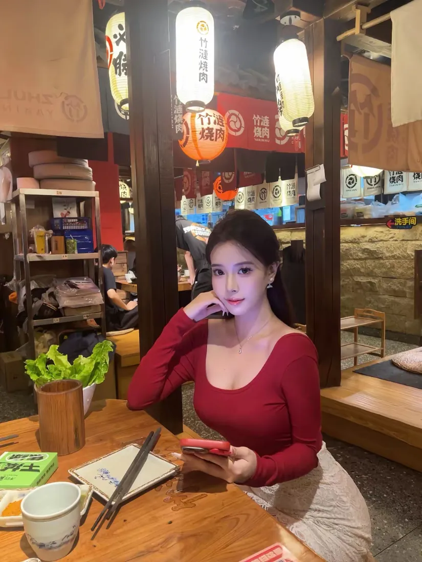 红衣长发女子坐在日式居酒屋餐桌旁，手持手机，手肘撑桌托腮。