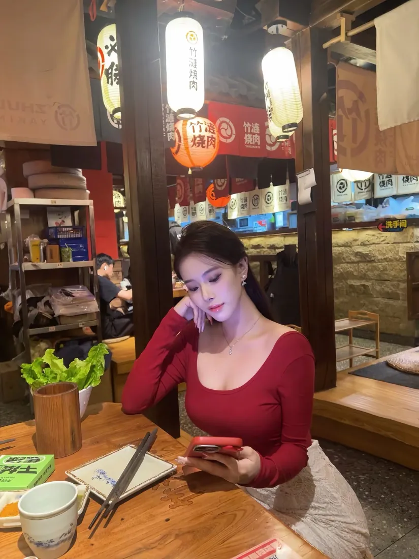 红衣长发女子坐在日式居酒屋餐桌旁，低头休闲看手机
