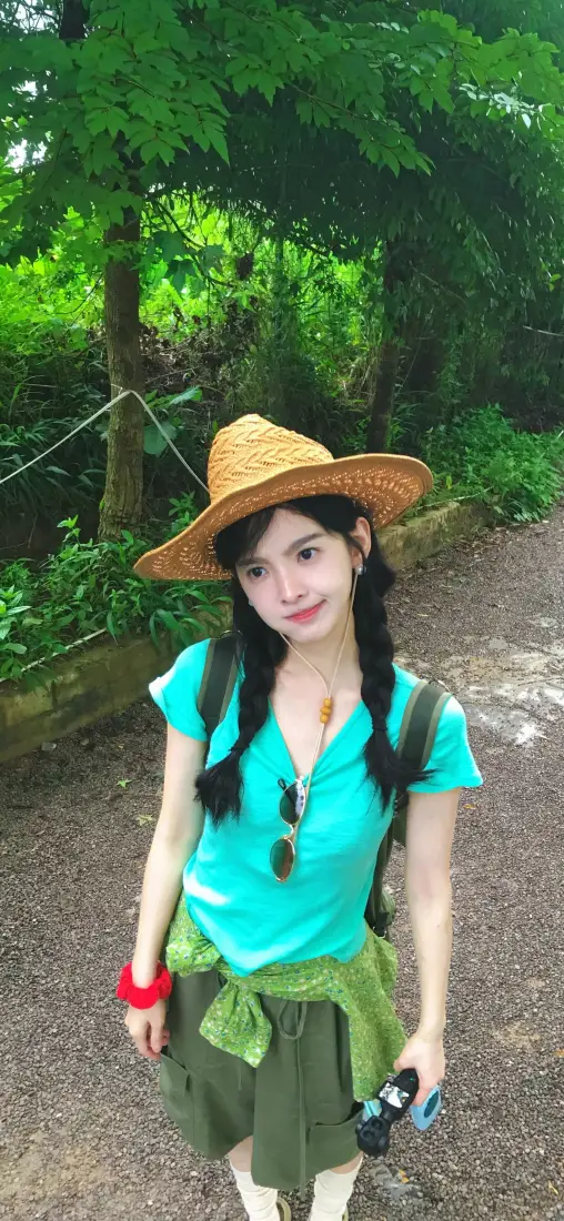 田曦薇戴草帽穿青绿色上衣，站在林间碎石步道上，手持相机微笑。