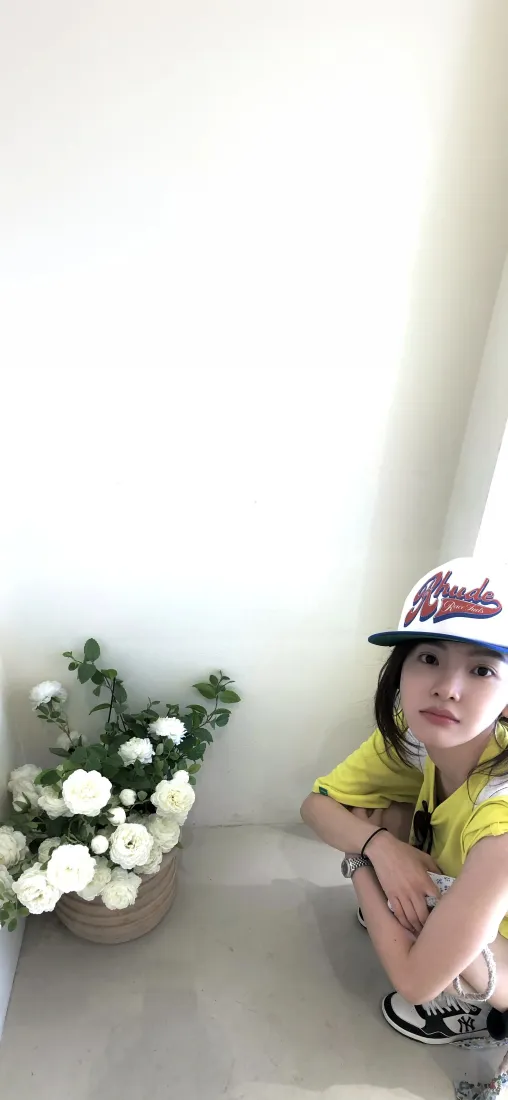 戴棒球帽穿黄色上衣的年轻女性蹲在室内白墙角落，身旁摆放白色花束盆栽。