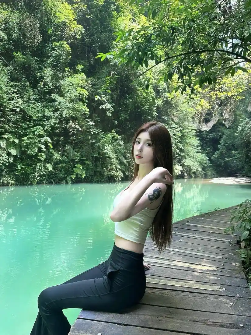 长发女子坐在林间碧水旁的木质栈道上，身着白色吊带与黑色长裤。