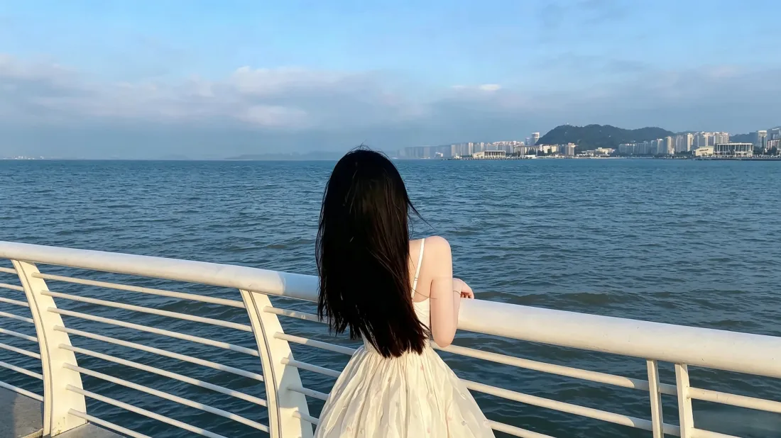 白裙长发少女背对镜头倚靠海边栏杆，眺望蓝海与远处城市岸线。