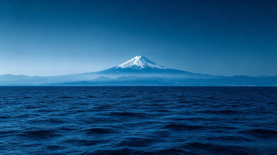 辽阔蓝色海面之上，远处云雾间矗立着覆雪的富士山，上方是澄澈蓝天。