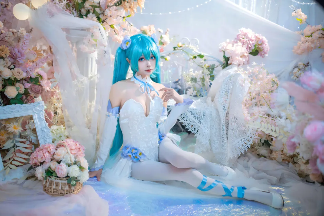 蓝发双马尾初音未来coser身着白裙白丝，坐于粉白花簇环绕的梦幻场景中