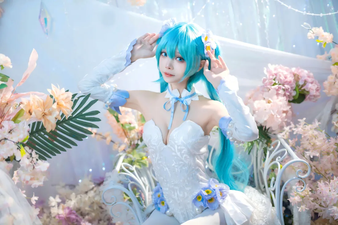 蓝发初音未来coser身着白裙，坐于花丛环绕的白色铁艺椅上，双手扶头。