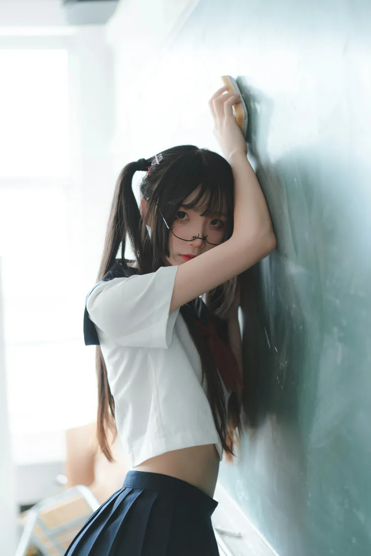 教室黑板前，双马尾戴眼镜少女穿露腰水手服倚墙摆拍