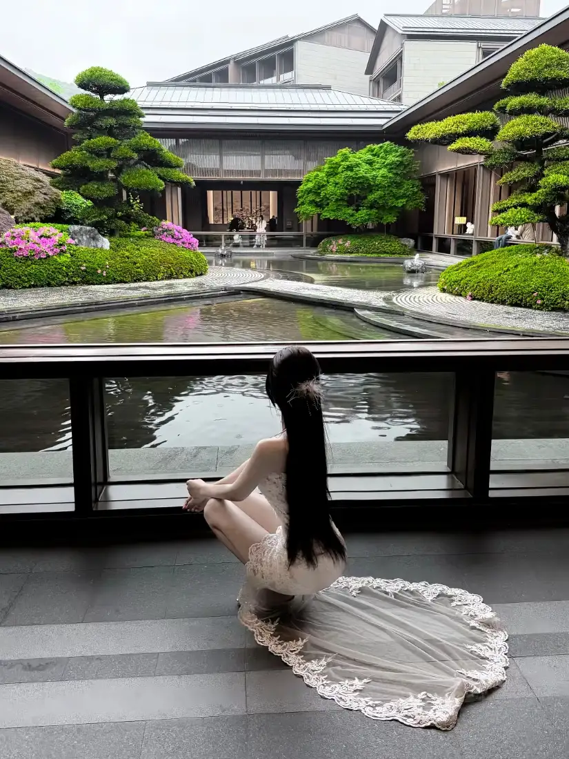 长发白衣女子蹲在中式庭院水景旁的阳台，背对镜头望向庭院池塘与绿植。