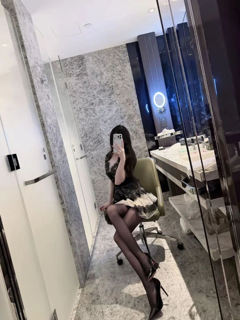 长发女子坐办公椅对镜自拍，着黑丝高跟与短裙，背景为大理石卫浴空间。