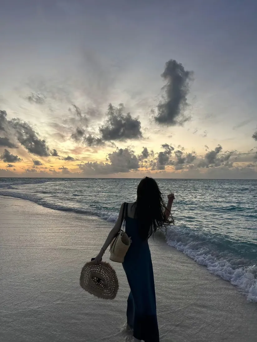 日落黄昏的海边沙滩上，穿深色长裙的女生背对镜头，手持草帽望向海面。