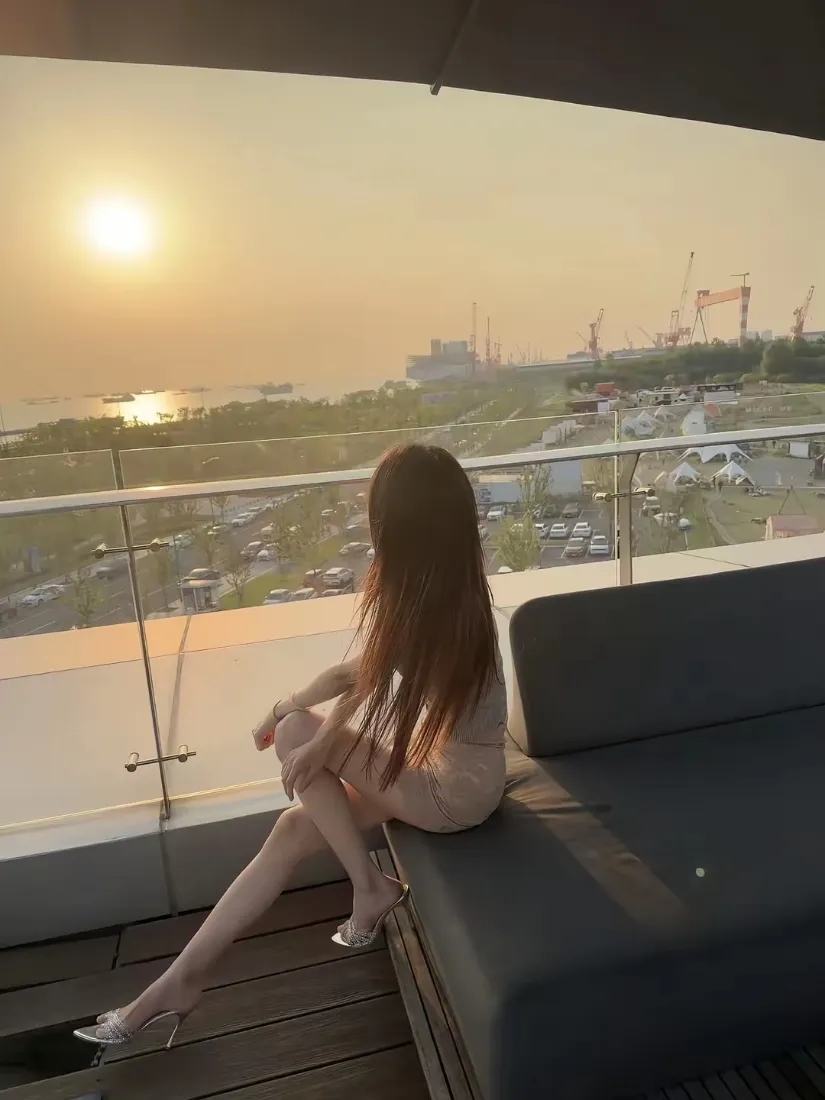 长发女子坐露台沙发上，背对镜头望向黄昏日落与远处城市景观
