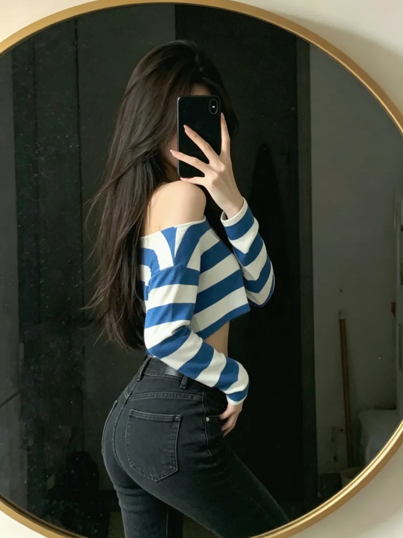 长发女子在圆形金框镜前对镜自拍，穿蓝白条纹露肩短上衣与黑色紧身牛仔裤。