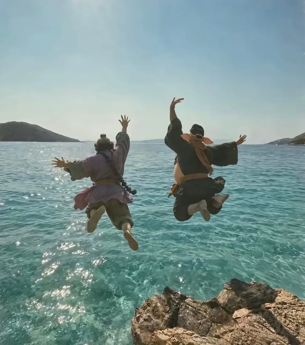 两名cos西游角色的男子背对镜头从礁石跃向碧海蓝天，姿态搞怪。