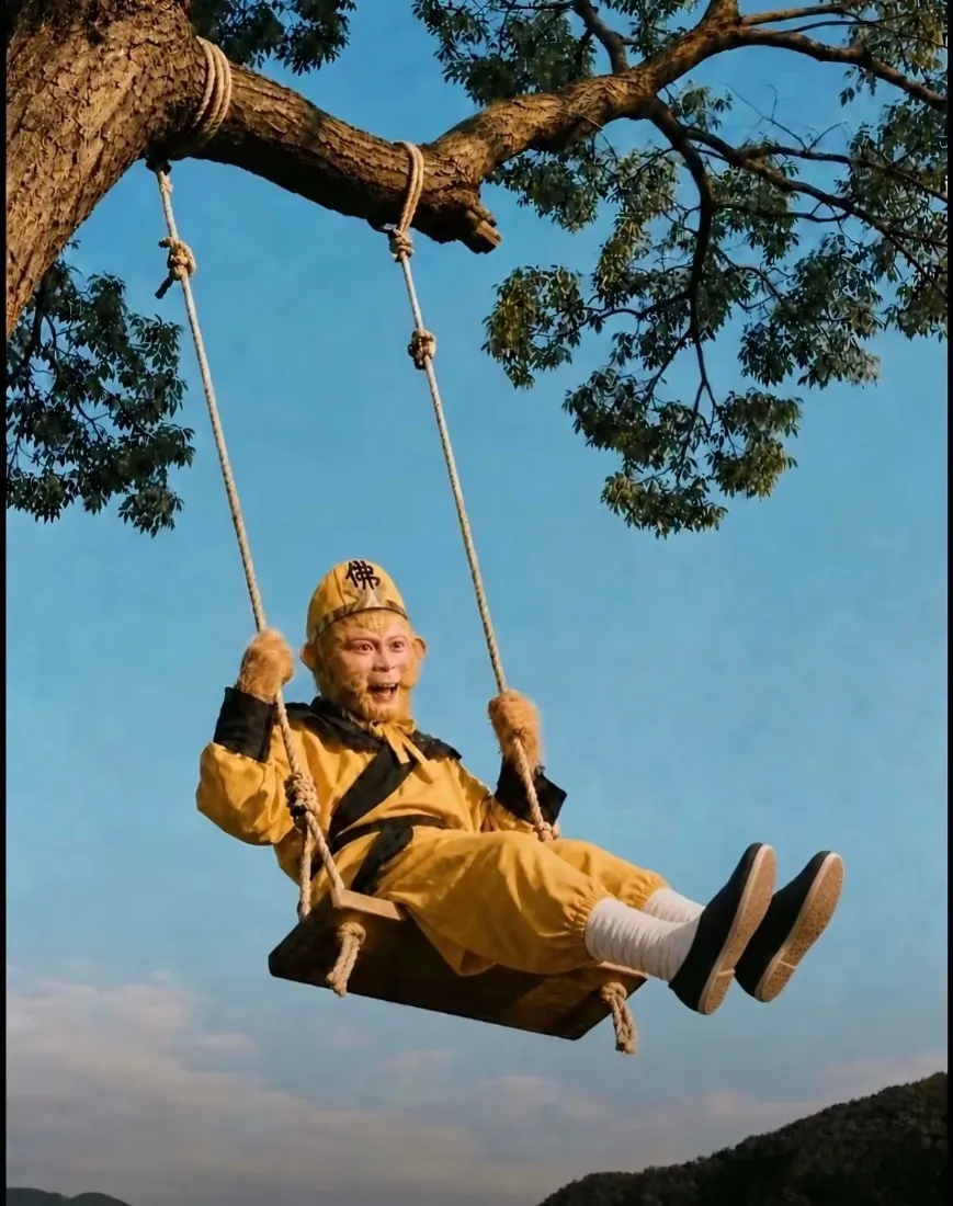 孙悟空装扮的人坐在大树枝悬挂的秋千上，背景是晴朗蓝天与远山。