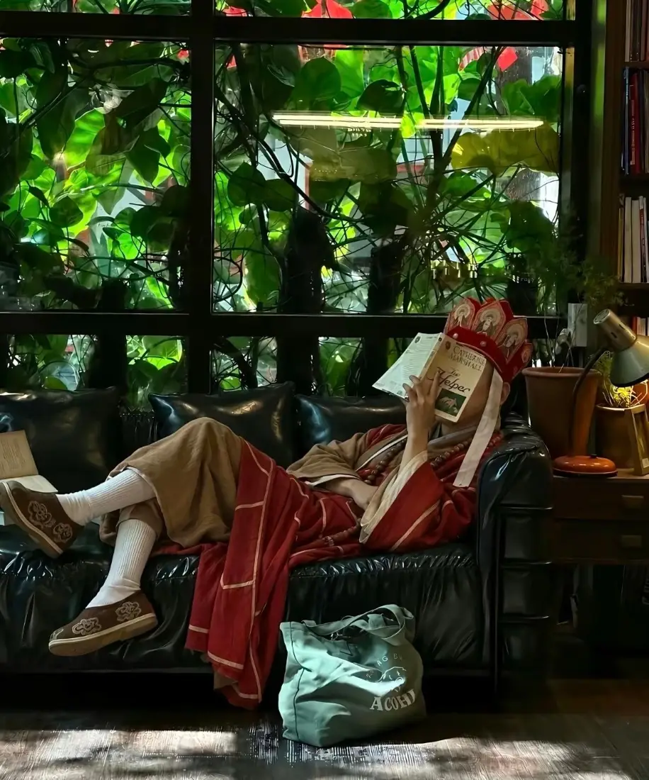 唐僧cosplay者躺卧在黑色皮沙发上，用书遮挡面部，窗边绿植环绕。