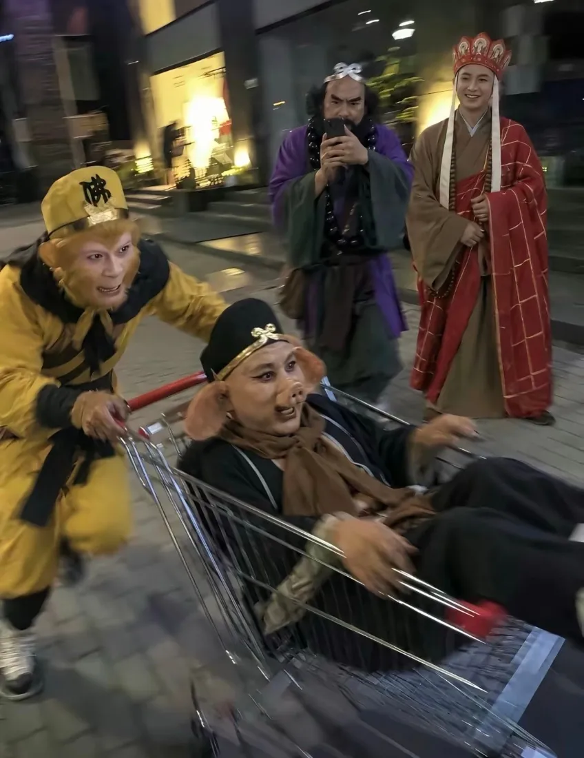 夜晚街头，西游记师徒四人cosplay，孙悟空推购物车载着猪八戒，沙僧持手机、唐僧随行。