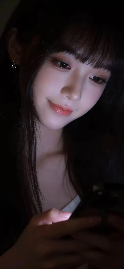 暗调环境中长发齐刘海女生低头浅笑，手持发光手机，氛围温柔朦胧