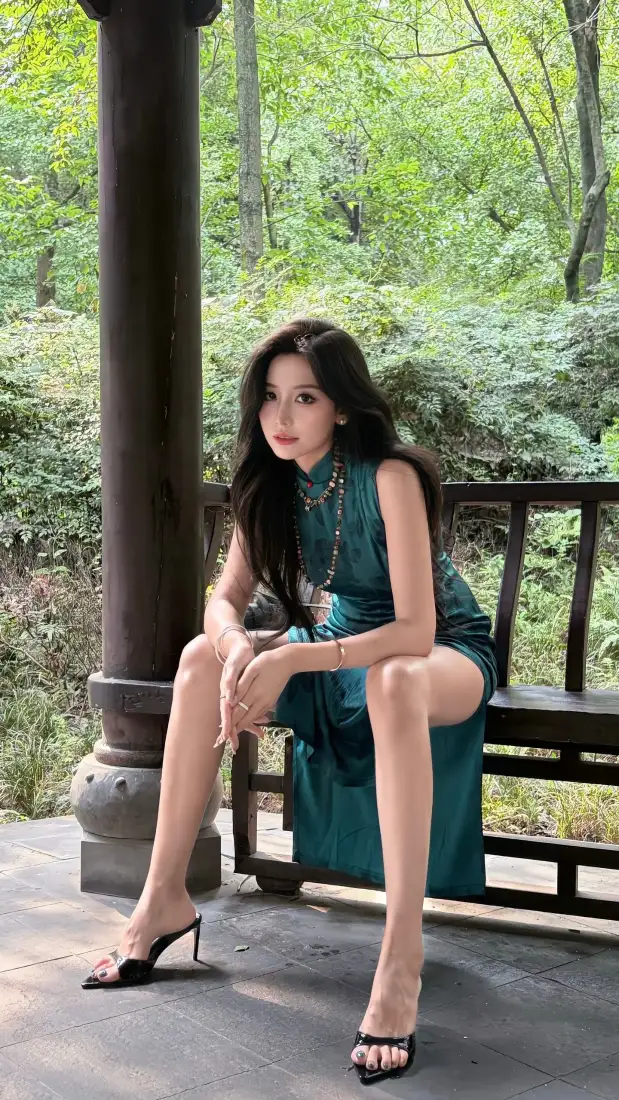 黑长直女子身着墨绿色无袖旗袍，坐于林间古亭长椅上。
